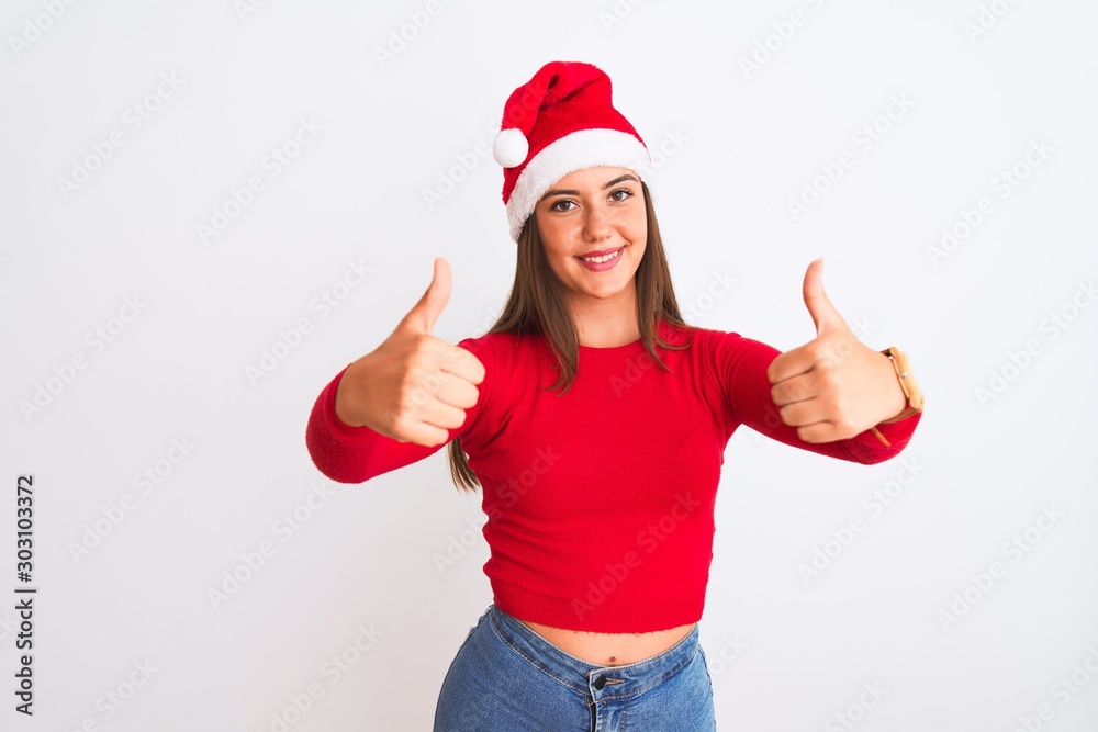 Young beautiful girl wearing Christmas Santa hat standing over isolated white background approving doing positive gesture with hand, thumbs up smiling and happy for success. Winner gesture. Young beautiful girl wearing Christmas Santa hat standing over isolated white background approving doing positive gesture with hand, thumbs up smiling and happy for success. Winner gesture.