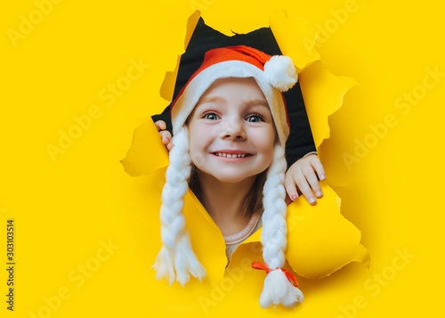 Waiting for a Christmas gift. A beautiful little smiling girl in a red Santa hat with pigtail looks out of a hole in yellow pastel paper. Christmas and New Year concept. Copy space. Torn background.