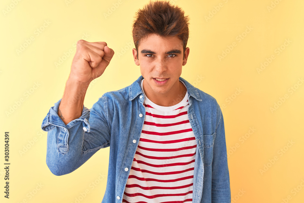 Fototapeta premium Young handsome man wearing striped t-shirt and denim shirt over isolated yellow background angry and mad raising fist frustrated and furious while shouting with anger. Rage and aggressive concept.