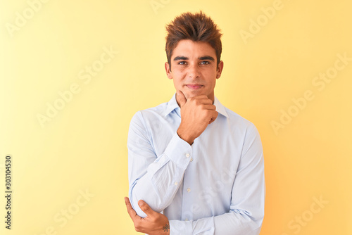 Young handsome businessman wearing elegant shirt over isolated yellow background looking confident at the camera with smile with crossed arms and hand raised on chin. Thinking positive.