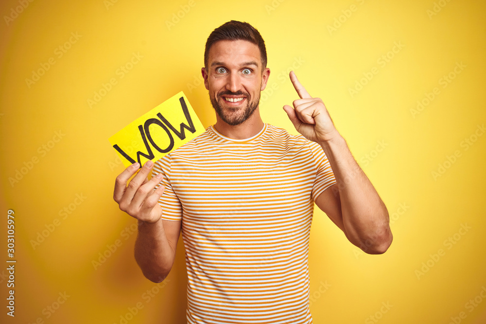 Young man holding wow text banner over yellow isolated background surprised with an idea or question pointing finger with happy face, number one Young man holding wow text banner over yellow isolated background surprised with an idea or question pointing finger with happy face, number one
