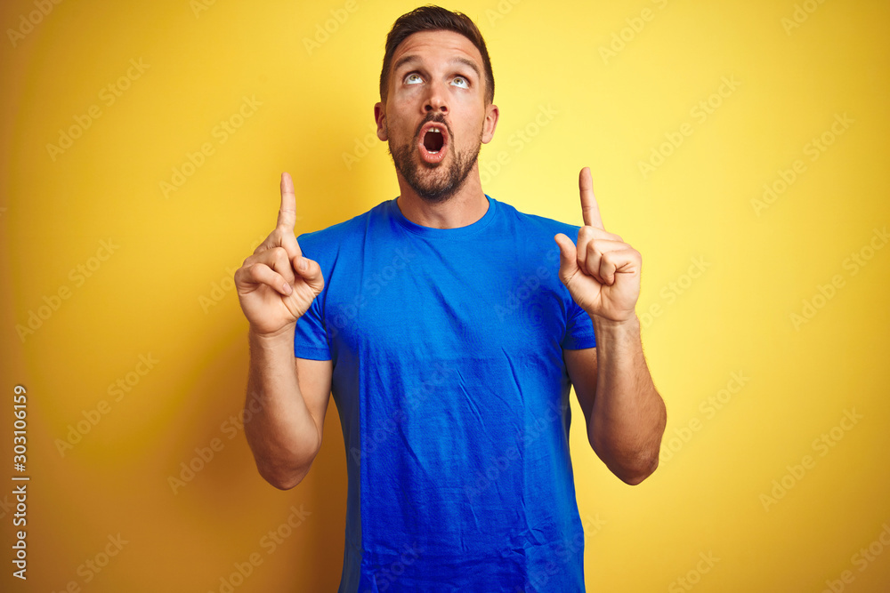 Young handsome man wearing casual blue t-shirt over yellow isolated background amazed and surprised looking up and pointing with fingers and raised arms. Young handsome man wearing casual blue t-shirt over yellow isolated background amazed and surprised looking up and pointing with fingers and raised arms.