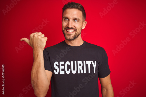 Young safeguard man wearing security uniform over red isolated background smiling with happy face looking and pointing to the side with thumb up.