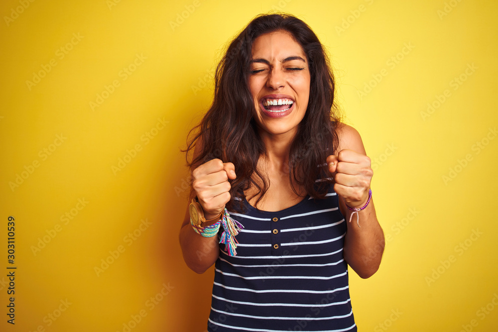 Young beautiful woman wearing striped t-shirt standing over isolated yellow background excited for success with arms raised and eyes closed celebrating victory smiling. Winner concept. Young beautiful woman wearing striped t-shirt standing over isolated yellow background excited for success with arms raised and eyes closed celebrating victory smiling. Winner concept.