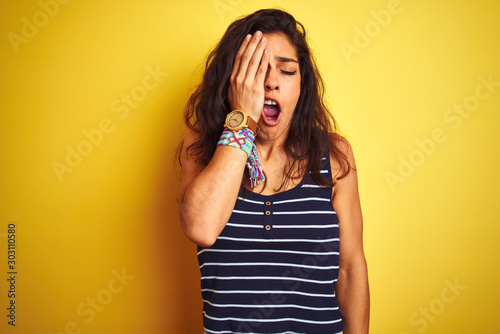 Young beautiful woman wearing striped t-shirt standing over isolated yellow background Yawning tired covering half face, eye and mouth with hand. Face hurts in pain.