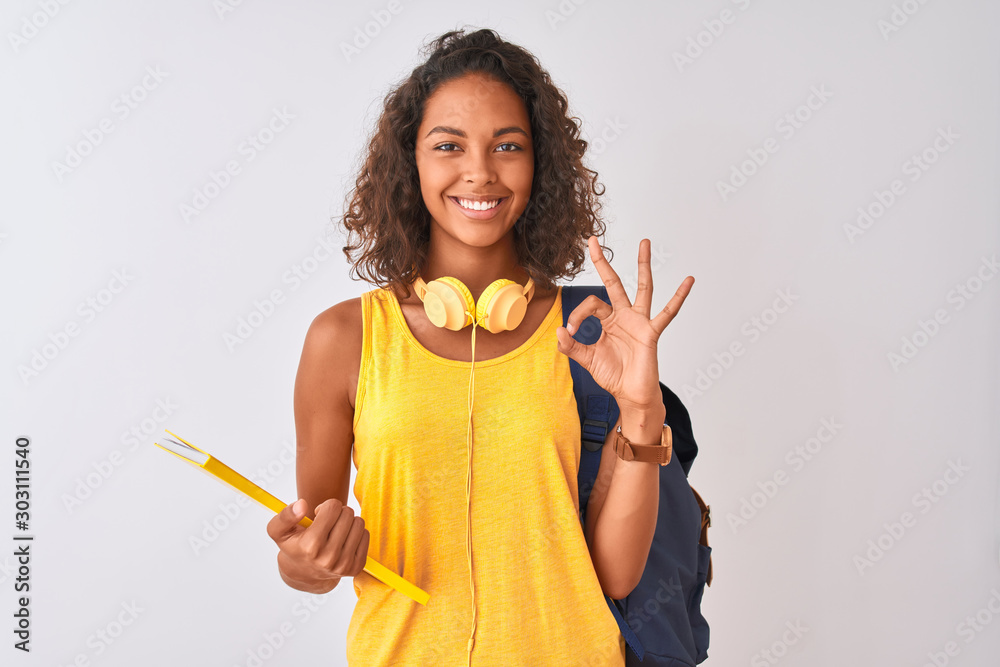 Brazilian student woman wearing backpack holding notebook over isolated white background doing ok sign with fingers, excellent symbol Brazilian student woman wearing backpack holding notebook over isolated white background doing ok sign with fingers, excellent symbol