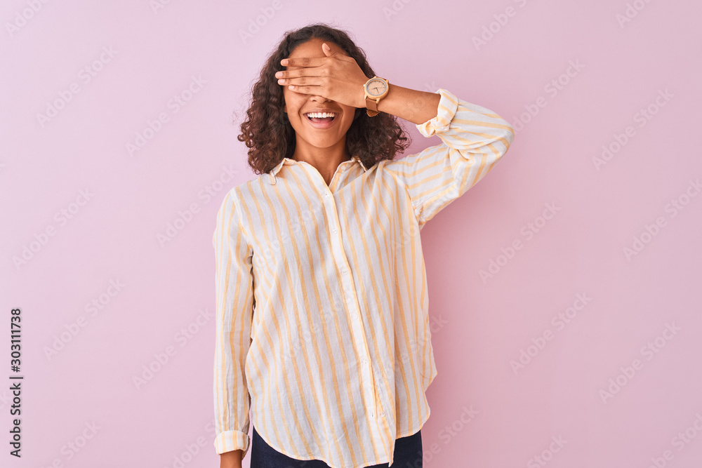 Young brazilian woman wearing striped shirt standing over isolated pink background smiling and laughing with hand on face covering eyes for surprise. Blind concept.