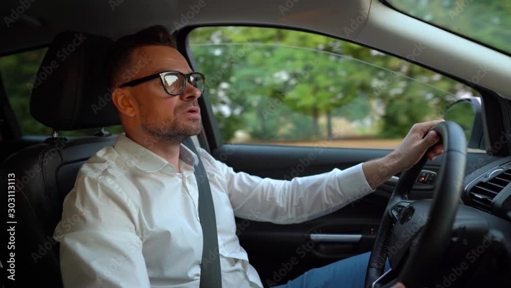 Bearded man in glasses and white shirt driving a car in sunny weather and swears at others behind the scenes Bearded man in glasses and white shirt driving a car in sunny weather and swears at others behind the scenes