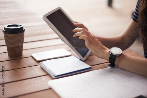 Women's hands with neat manicure and large wristwatch, work with gadgets, tablet, smartphone, papers on a wooden table.