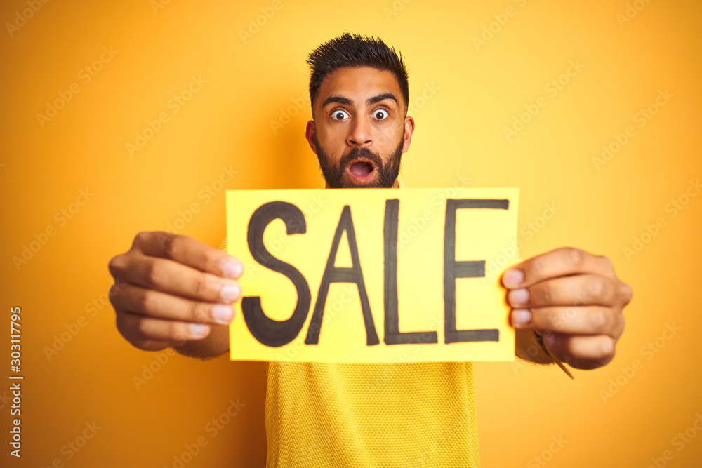 Young arab indian hispanic man holding sale banner over isolated yellow background scared in shock with a surprise face, afraid and excited with fear expression Young arab indian hispanic man holding sale banner over isolated yellow background scared in shock with a surprise face, afraid and excited with fear expression