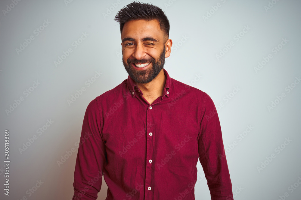 Obraz premium Young indian man wearing red elegant shirt standing over isolated grey background winking looking at the camera with sexy expression, cheerful and happy face.