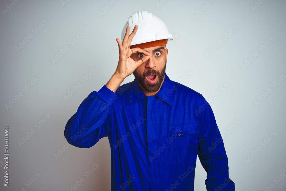 Handsome indian worker man wearing uniform and helmet over isolated white background doing ok gesture shocked with surprised face, eye looking through fingers. Unbelieving expression.
