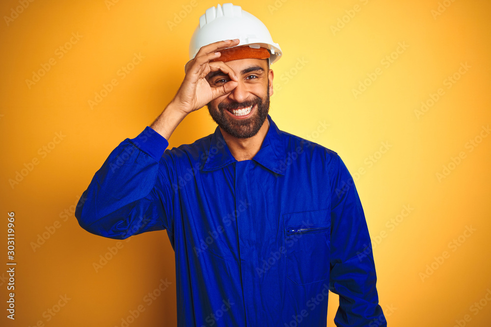 Handsome indian worker man wearing uniform and helmet over isolated yellow background doing ok gesture with hand smiling, eye looking through fingers with happy face.