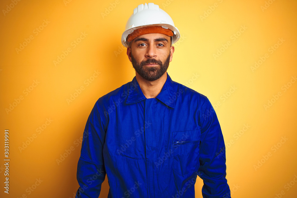 Handsome indian worker man wearing uniform and helmet over isolated yellow background with serious expression on face. Simple and natural looking at the camera.