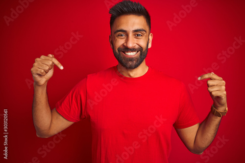 Quadro su tela Young handsome indian man wearing t-shirt over isolated red background looking confident with smile on face, pointing oneself with fingers proud and happy