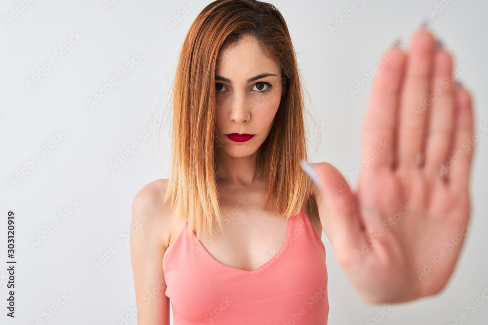 Beautiful redhead woman wearing casual pink t-shirt standing over isolated white background with open hand doing stop sign with serious and confident expression, defense gesture Beautiful redhead woman wearing casual pink t-shirt standing over isolated white background with open hand doing stop sign with serious and confident expression, defense gesture