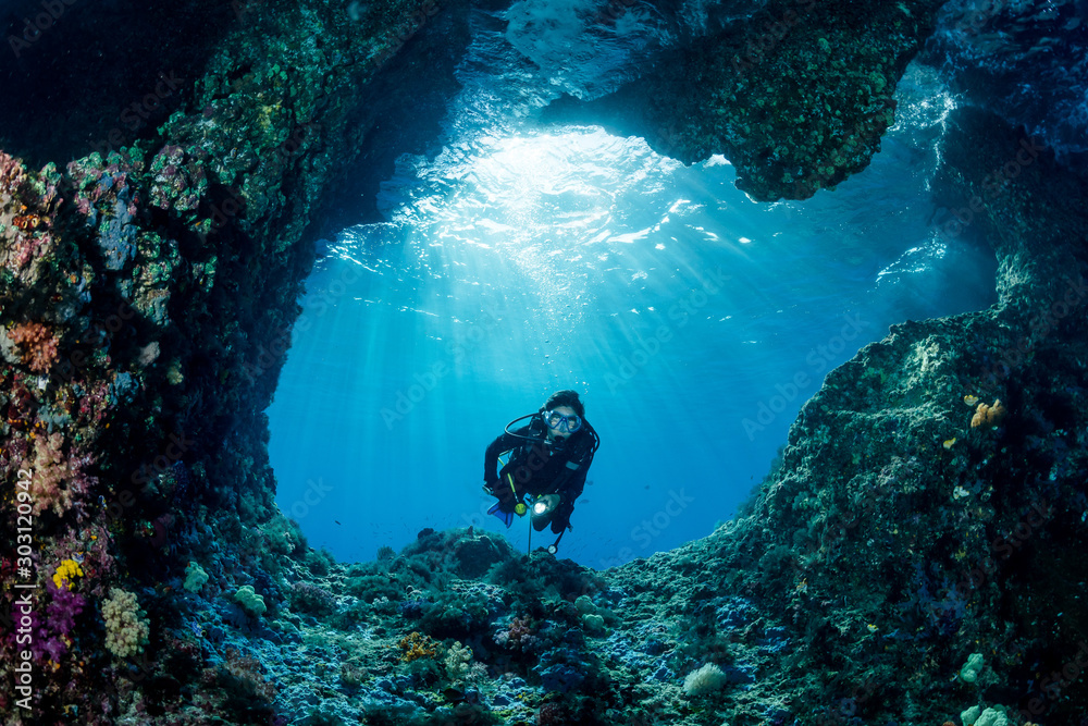 woman diver underwater at the entrance of a cave with sunrays - obrazy, fototapety, plakaty 