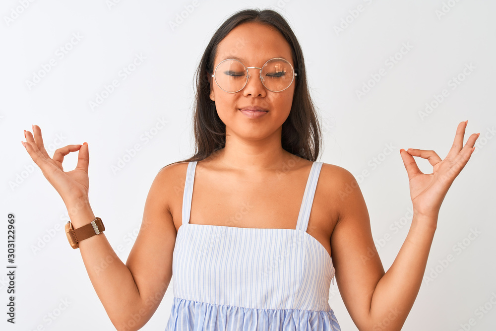 Young chinese woman wearing striped dress and glasses over isolated white background relax and smiling with eyes closed doing meditation gesture with fingers. Yoga concept. Young chinese woman wearing striped dress and glasses over isolated white background relax and smiling with eyes closed doing meditation gesture with fingers. Yoga concept.