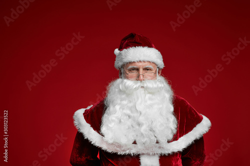 Portrait of senior man wearing Santa costume with white beard looking at camera isolated on red background