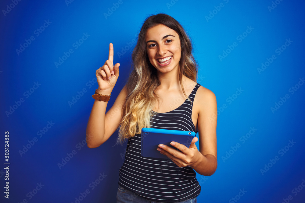 Young beautiful woman using touchpad tablet over blue isolated background surprised with an idea or question pointing finger with happy face, number one Young beautiful woman using touchpad tablet over blue isolated background surprised with an idea or question pointing finger with happy face, number one