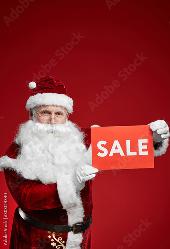 Portrait of Santa Claus in costume and with white beard holding sale sign against the red background