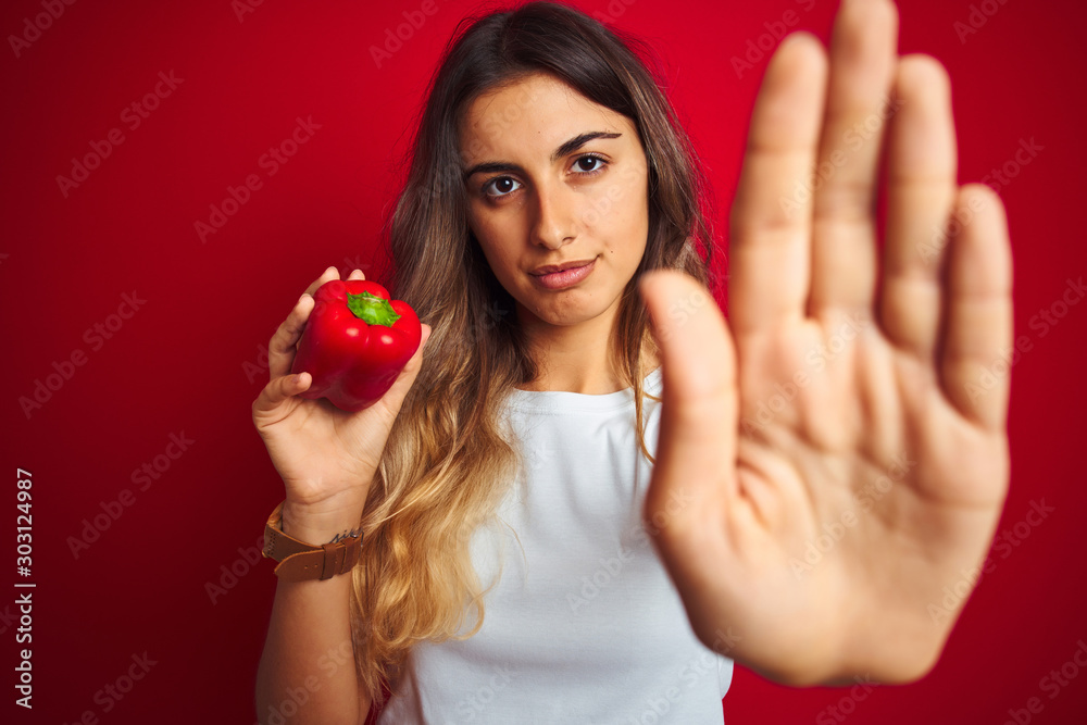 Young beautiful woman holding pepper over red isolated background with open hand doing stop sign with serious and confident expression, defense gesture Young beautiful woman holding pepper over red isolated background with open hand doing stop sign with serious and confident expression, defense gesture