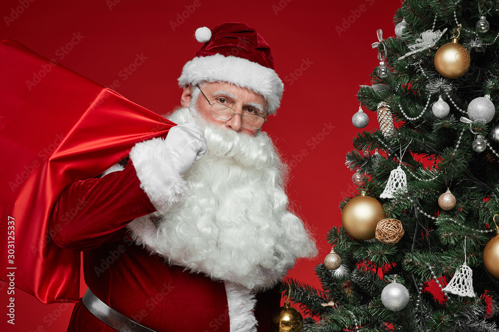 Portrait of Santa Claus with big sack behind his back carrying presents for Christmas isolated on red background Portrait of Santa Claus with big sack behind his back carrying presents for Christmas isolated on red background