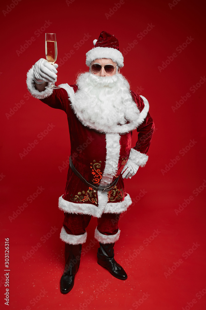Portrait of Santa Claus in sunglasses standing and holding glass of champagne in his hand he celebrating Christmas holidays Portrait of Santa Claus in sunglasses standing and holding glass of champagne in his hand he celebrating Christmas holidays