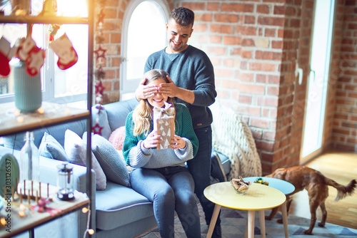 Young beautiful couple smiling happy and confident. Man covering eyes of woman surpise her with gift around christmas decoration at home © Krakenimages.com