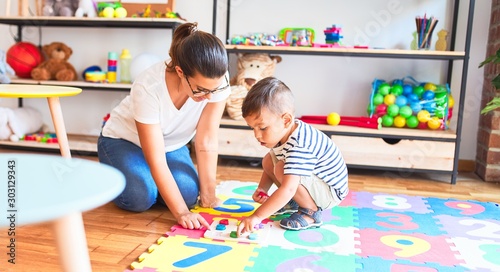 Beautiful teacher and toddler boy sitting on puzzle playing with numbers at kindergarten