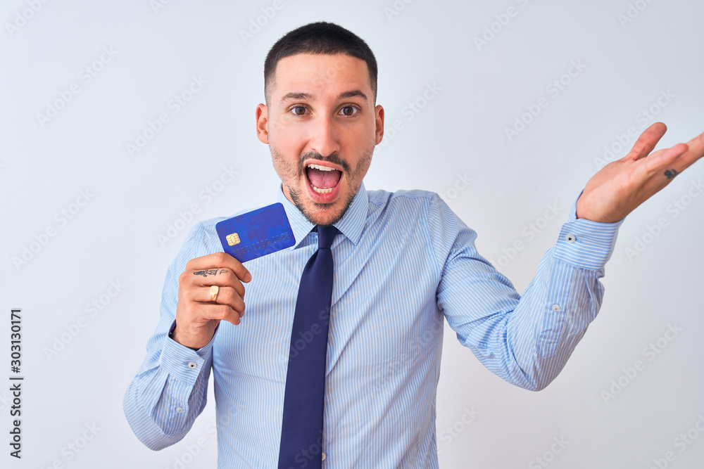 Young business man holding credit card over isolated background very happy and excited, winner expression celebrating victory screaming with big smile and raised hands Young business man holding credit card over isolated background very happy and excited, winner expression celebrating victory screaming with big smile and raised hands