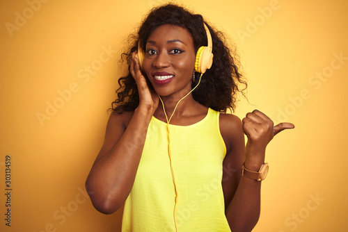 African american woman listening to music using headphones over isolated yellow background pointing and showing with thumb up to the side with happy face smiling