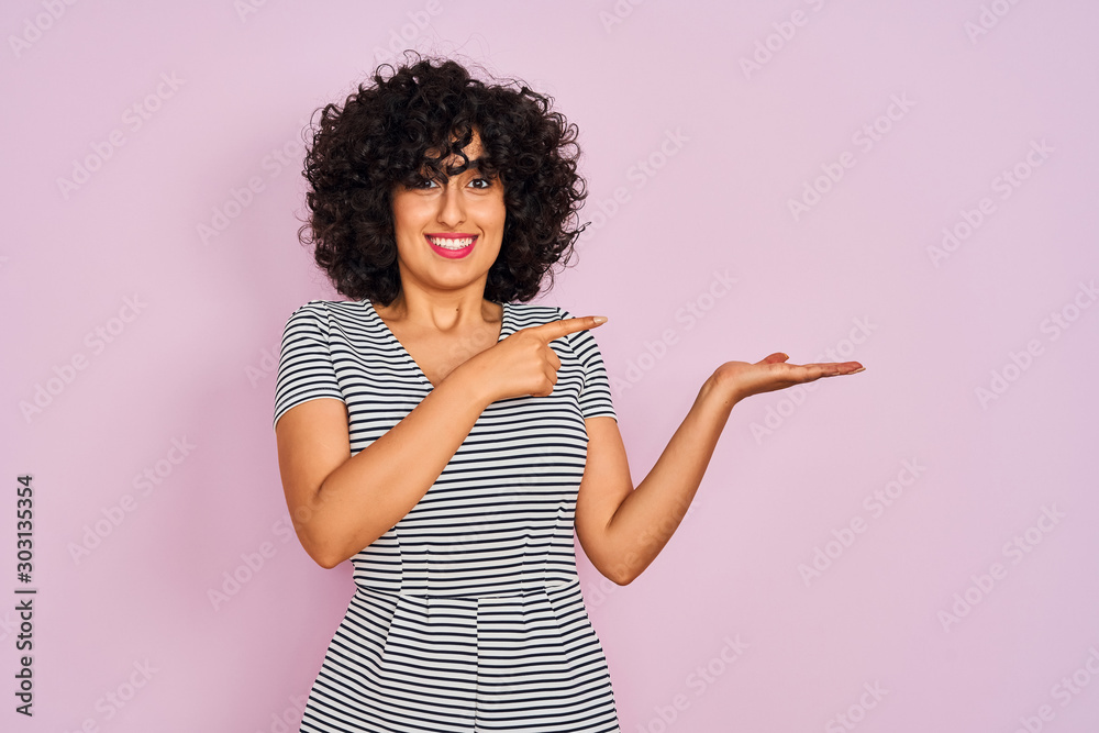 Young arab woman with curly hair wearing striped dress over isolated pink background amazed and smiling to the camera while presenting with hand and pointing with finger. Young arab woman with curly hair wearing striped dress over isolated pink background amazed and smiling to the camera while presenting with hand and pointing with finger.