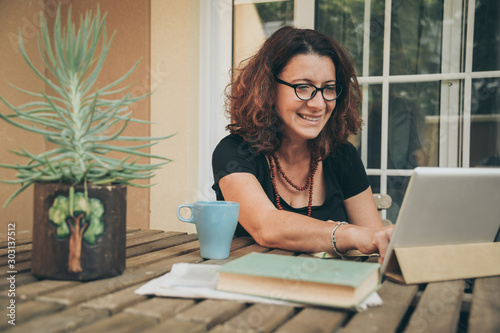 Middle aged female studying at home with books, newspaper and digital tablet pad. Woman reading a book and watching video online on new tech device. Education, modern lifestyle and leisure concept. photo