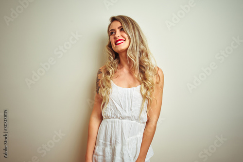 Young beautiful woman wearing casual dress standing over white isolated background looking away to side with smile on face, natural expression. Laughing confident.