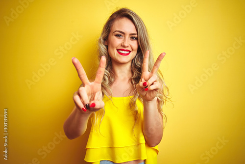 Young attactive woman wearing t-shirt standing over yellow isolated background smiling looking to the camera showing fingers doing victory sign. Number two.