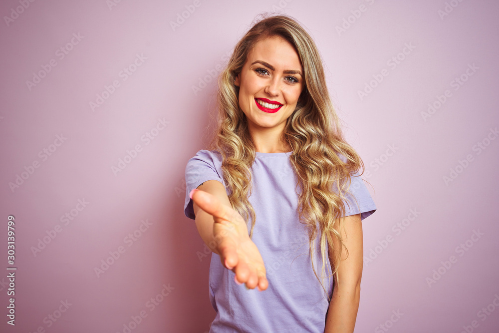 Young beautiful woman wearing purple t-shirt standing over pink isolated background smiling friendly offering handshake as greeting and welcoming. Successful business. Young beautiful woman wearing purple t-shirt standing over pink isolated background smiling friendly offering handshake as greeting and welcoming. Successful business.