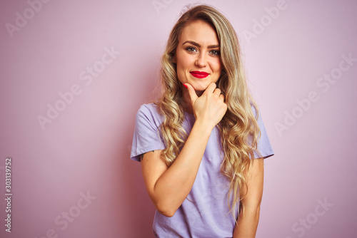 Young beautiful woman wearing purple t-shirt standing over pink isolated background looking confident at the camera with smile with crossed arms and hand raised on chin. Thinking positive.