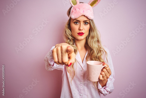 Young woman wearing pajama and mask drinking a cup of coffee over pink isolated background pointing with finger to the camera and to you, hand sign, positive and confident gesture from the front