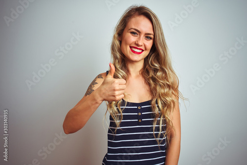 Young beautiful woman wearing stripes t-shirt standing over white isolated background doing happy thumbs up gesture with hand. Approving expression looking at the camera showing success.