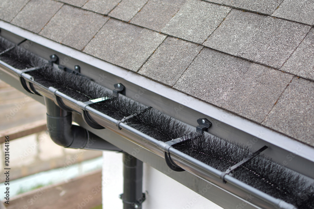 Obraz premium Plastic guard brush in new dark grey plastic rain gutter on asphalt shingles roof.