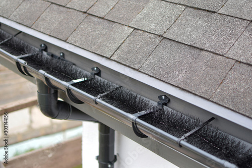 Plastic guard brush in new dark grey plastic rain gutter on asphalt shingles roof. © Inga