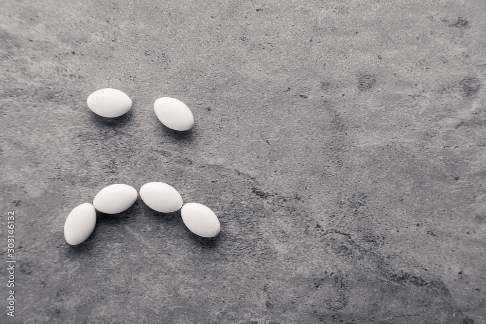 Sad smile from scattered white medicine vitamins, pills, drugs on gray background. White food diet supplement. Black and white photography. Top view Sad smile from scattered white medicine vitamins, pills, drugs on gray background. White food diet supplement. Black and white photography. Top view