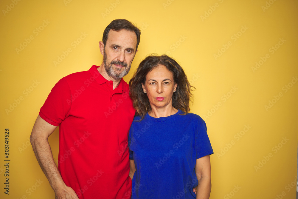 Beautiful middle age couple together standing over isolated yellow background with serious expression on face. Simple and natural looking at the camera. Beautiful middle age couple together standing over isolated yellow background with serious expression on face. Simple and natural looking at the camera.
