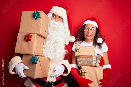 Middle age couple wearing Santa costume holding tower of gifts over isolated red background smiling looking to the side and staring away thinking.