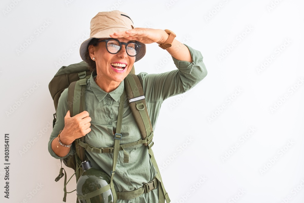 Middle age hiker woman wearing backpack canteen hat glasses over isolated white background very happy and smiling looking far away with hand over head. Searching concept.