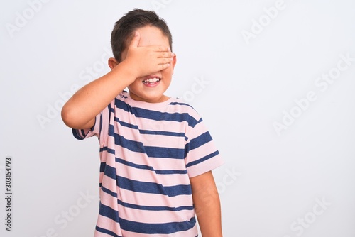 Beautiful kid boy wearing casual striped t-shirt standing over isolated white background smiling and laughing with hand on face covering eyes for surprise. Blind concept.