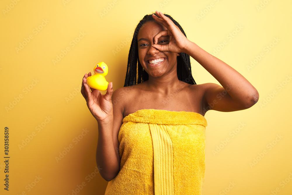 African american woman wearing shower towel holding toy duck over isolated yellow background with happy face smiling doing ok sign with hand on eye looking through fingers African american woman wearing shower towel holding toy duck over isolated yellow background with happy face smiling doing ok sign with hand on eye looking through fingers