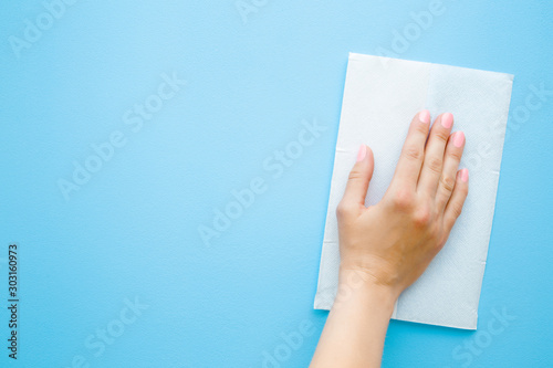 Woman hand wiping pastel blue desk with white paper napkin. General or regular cleanup. Closeup. Empty place for text or logo. Top down view.