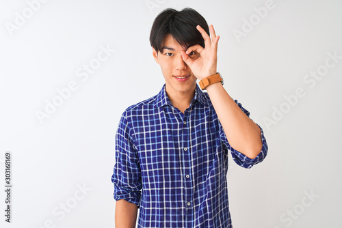 Young chinese man wearing casual blue shirt standing over isolated white background doing ok gesture with hand smiling, eye looking through fingers with happy face.
